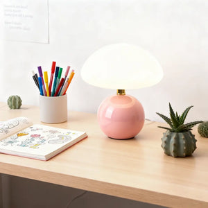 Abajur cogumelo rosa em uma escrivaninha de madeira, ao lado de lápis de cor e cactos. A luminária de mesa de cerâmica inspira criatividade e personalidade
