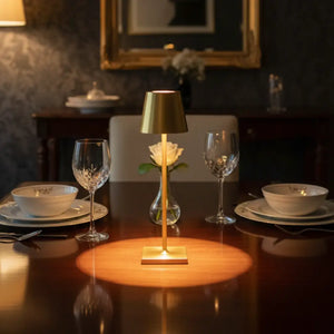 Luminária de mesa dourada no centro de uma mesa de jantar de madeira, com taças de vinho e louças, criando uma atmosfera sofisticada e íntima