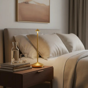Luminária de mesa portátil dourada sobre uma mesa de cabeceira em um quarto de luxo, ao lado de livros e uma jarra de vidro, ideal para leitura e relaxamento