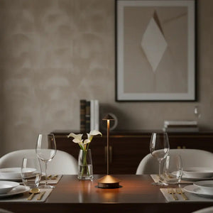 Luminária de mesa sem fio com design minimalista, usada para iluminar uma mesa de jantar de madeira escura com taças de vinho e louças, criando uma atmosfera aconchegante