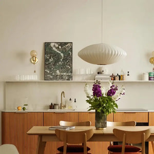 Luminária pendente japonesa oval em seda branca pendurada sobre uma mesa de jantar de madeira clara em uma cozinha moderna com bancada de mármore e armários inferiores de madeira ripada.