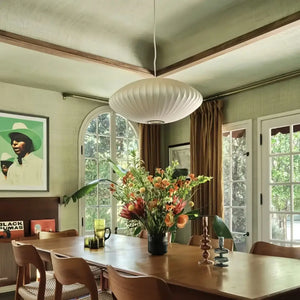 Luminária pendente japonesa oval em seda branca sobre uma mesa de jantar de madeira maciça em uma sala rústica com teto verde texturizado, vigas de madeira e uma grande janela em arco