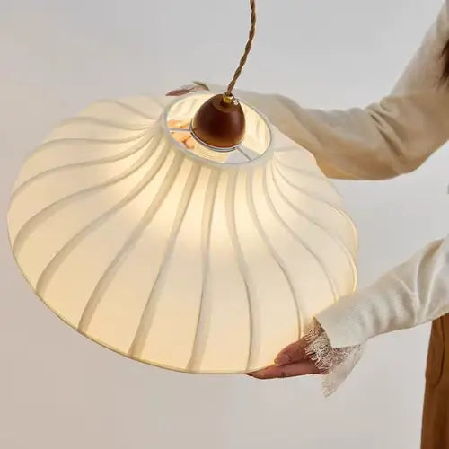 Mão segurando a Luminaria Pendente Moderno Minimalista Madeira Tecido Radiant — detalhe das pregas, cabo trançado dourado, topo walnut
