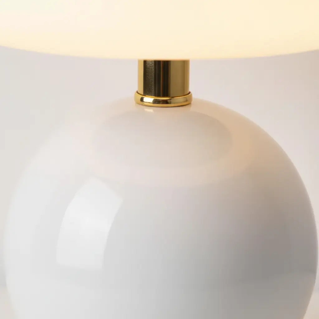 Close-up detalhado do abajur de cerâmica branco, mostrando o brilho da base esférica e o conector de metal dourado que une a cúpula e a base. Realça o acabamento premium