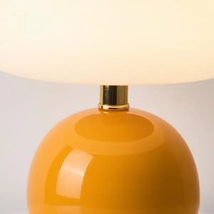 Close-up detalhado do abajur cogumelo laranja, destacando a base de cerâmica brilhante e o conector de metal dourado entre a base e a cúpula. Revela a qualidade premium dos materiais