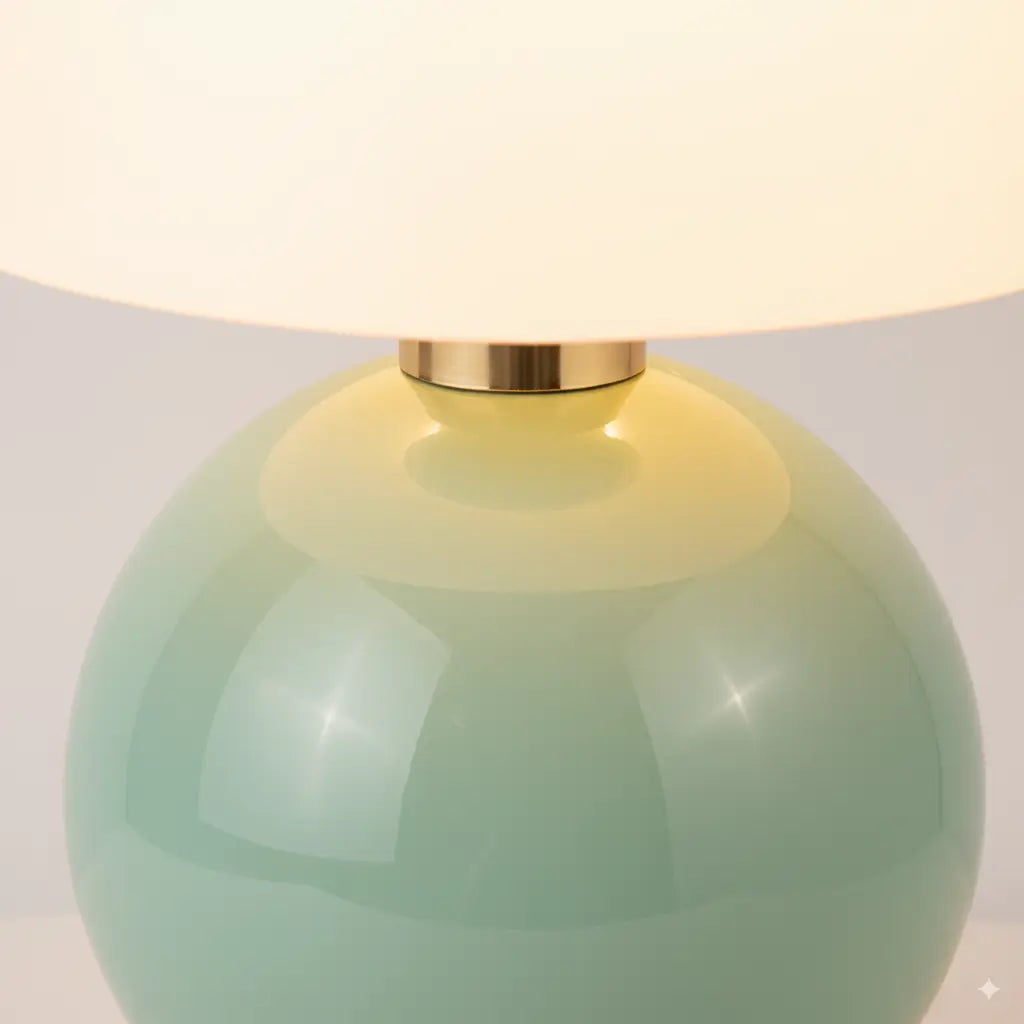 Close-up da base do abajur verde menta, com luz suave criando um reflexo que destaca o acabamento brilhante da cerâmica e o conector dourado polido