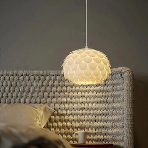 Close-up da Luminária Pendente Pétala acesa sobre uma mesa de cabeceira, destacando a textura fosca das camadas e a luz suave contra uma cabeceira de tricô