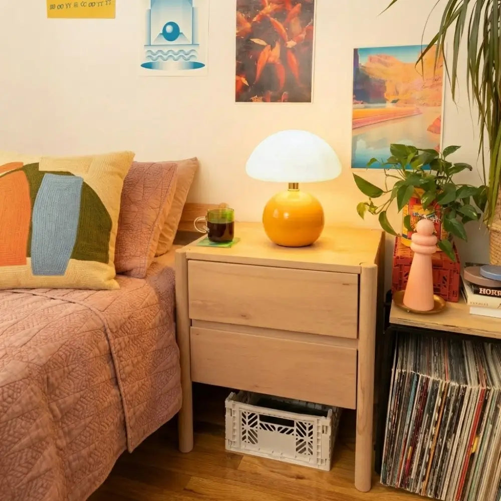 Luminária Cogumelo Laranja acesa sobre uma mesa de cabeceira de madeira, decorando um quarto aconchegante com livros e plantas. Iluminação indireta para leitura