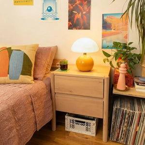 Luminária Cogumelo Laranja acesa sobre uma mesa de cabeceira de madeira, decorando um quarto aconchegante com livros e plantas. Iluminação indireta para leitura
