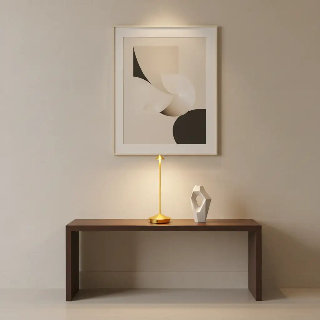Luminária de mesa dourada em um ambiente minimalista, posicionada sobre um console e sob uma obra de arte abstrata, mostrando seu uso como objeto de design