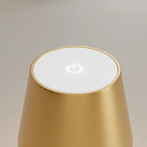 Close-up da parte superior da luminária de mesa dourada, mostrando o botão de toque que controla o liga/desliga e a intensidade da luz LED