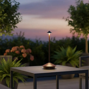 Luminária de mesa portátil na cor marrom em um ambiente externo, sobre uma mesa em uma varanda ou jardim, com luz suave no entardecer