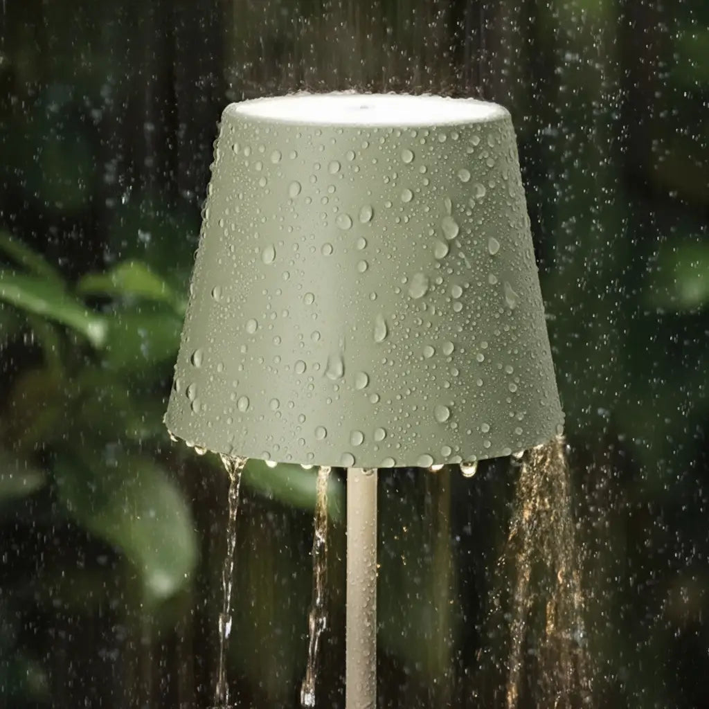 Luminária de mesa verde sendo usada em um ambiente externo, com gotas de água escorrendo sobre a cúpula, mostrando sua resistência para uso em jardins e varandas