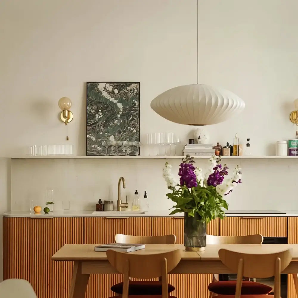 Luminária pendente japonesa oval em seda branca pendurada sobre uma mesa de jantar de madeira clara em uma cozinha moderna com bancada de mármore e armários inferiores de madeira ripada.