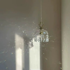 Luminária pendente de vidro facetado tipo cristal Marquise Radiantbr apagada, recebendo luz solar direta que projeta sombras e reflexos prismáticos na parede clara.