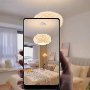 Mão segurando um smartphone que exibe uma simulação em realidade aumentada da Luminária Pendente Pétala instalada em um quarto moderno